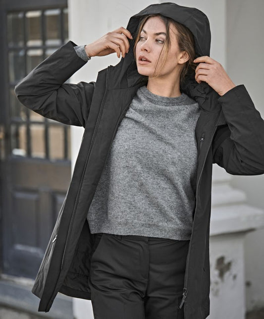 Damen All Weather Parka in in Schwarz mit geradem, längerem Schnitt bis über die Hüfte. Schlichtes Design mit hochstehendem Kragen und großer Kapuze, verdecktem Frontreißverschluss und zwei seitlichen Reißverschlusstaschen