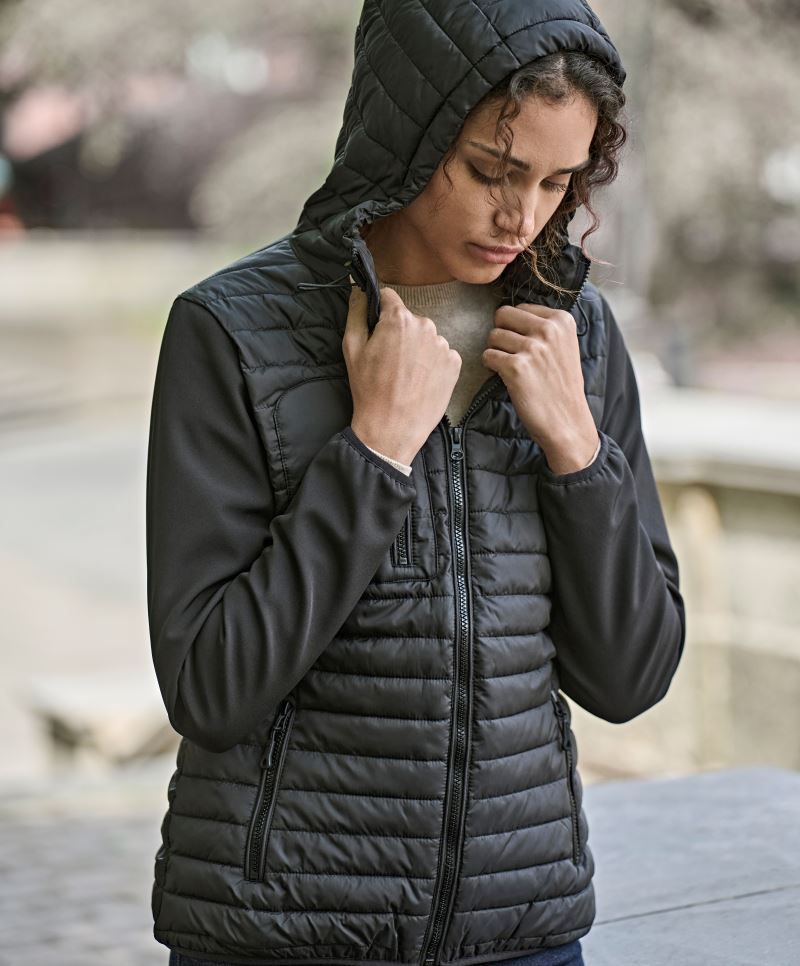 Damen Hooded Crossover Jacke in schwarz mit horizontal gesteppter Vorderseite und glatten Stretch-Ärmeln. Körpernah geschnitten mit Kapuze, hohem Kragen und durchgehendem Frontreißverschluss.