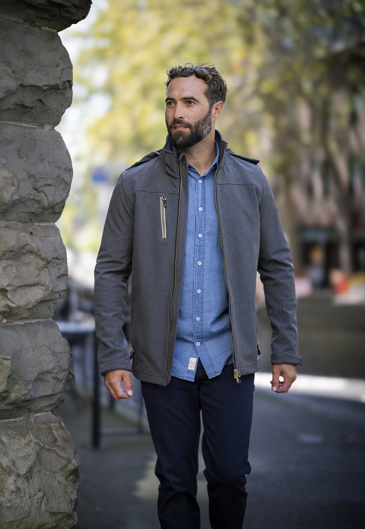 Hüftlange Softshelljacke für Herren mit Kapuze, hohem Stehkragen und verstellbaren Ärmelabschlüssen. Auffällig sind die Reißverschlusstasche auf der Brust und die beiden seitlichen Reißverschlusstaschen.
