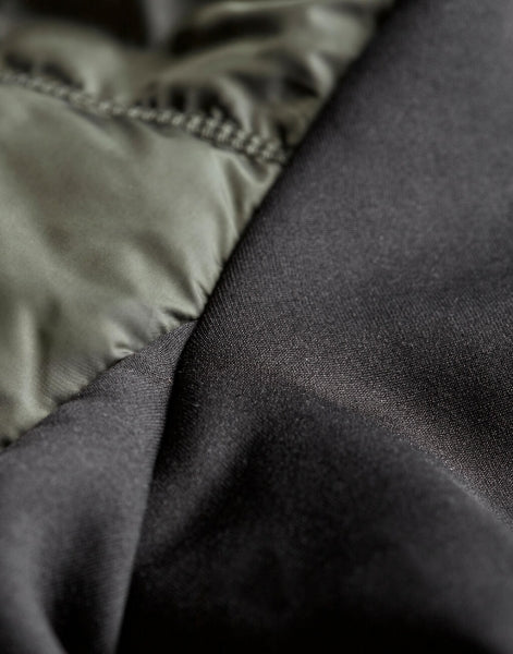 Herren Hybrid-Stretch-Jacke mit wattierter, quer gesteppter Vorderseite in Dunkelgrün, schwarzen Stretch-Ärmeln und seitlichen Einsätzen. Die Jacke ist körpernah geschnitten und hat einen hohen Stehkragen sowie einen durchgehenden Front Reißverschluss.
