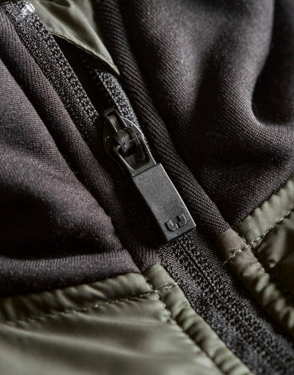 Herren Hybrid-Stretch-Jacke mit wattierter, quer gesteppter Vorderseite in Dunkelgrün, schwarzen Stretch-Ärmeln und seitlichen Einsätzen. Die Jacke ist körpernah geschnitten und hat einen hohen Stehkragen sowie einen durchgehenden Front Reißverschluss.