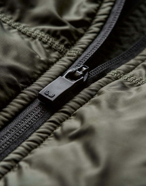 Herren Hybrid-Stretch-Weste mit wattierter, quer gesteppter Vorderseite in tiefen grün und seitlichen Einsätzen. Die Weste ist körpernah geschnitten und hat einen hohen Stehkragen sowie einen durchgehenden Front Reißverschluss.