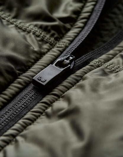 Herren Hybrid-Stretch-Weste mit wattierter, quer gesteppter Vorderseite in tiefen grün und seitlichen Einsätzen. Die Weste ist körpernah geschnitten und hat einen hohen Stehkragen sowie einen durchgehenden Front Reißverschluss.