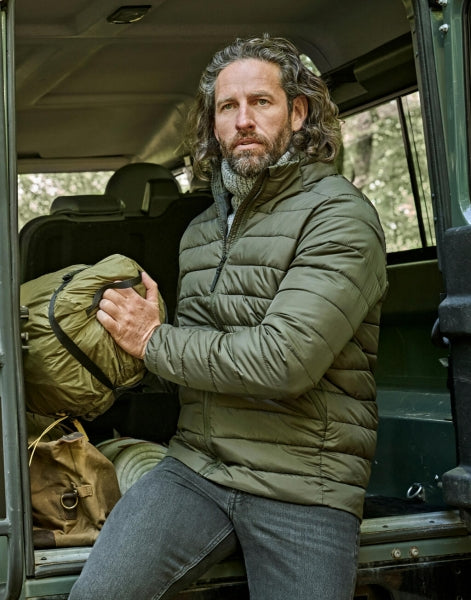 Ein Mann trägt eine Deep Green Lite Jacket mit schmaler, horizontaler Steppung und hohem Stehkragen. Die leicht wattierte Jacke reicht bis zur Hüfte und ist körpernah geschnitten. Zwei seitliche Taschen sind dezent in das Design integriert.
