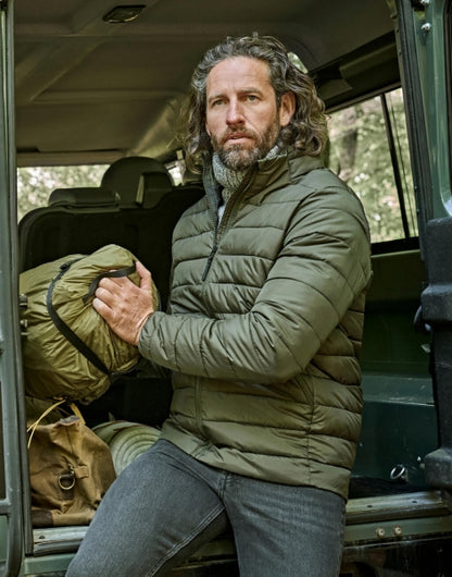 Ein Mann trägt eine Deep Green Lite Jacket mit schmaler, horizontaler Steppung und hohem Stehkragen. Die leicht wattierte Jacke reicht bis zur Hüfte und ist körpernah geschnitten. Zwei seitliche Taschen sind dezent in das Design integriert.
