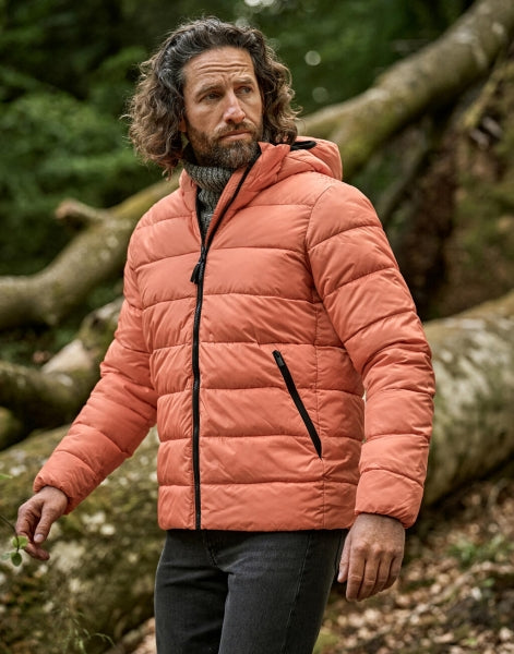 Ein Mann trägt eine Spicy Orange leichte Kapuzenjacke ist mit schmaler, horizontaler Steppung und hohem Stehkragen. Die leicht wattierte Jacke reicht bis zur Hüfte und ist körpernah geschnitten. Zwei seitliche Taschen sind dezent in das Design integriert.
