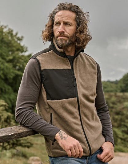 Mann trägt eine Mountain Fleece Bodywarmer in Clay/Black mit schwarzem Reißverschluss und dunklen Kontrastbesätzen an Brust und Schultern. Die hüftlange Weste ist körpernah geschnitten, hat einen hohen Kragen und seitliche Reißverschlusstaschen.