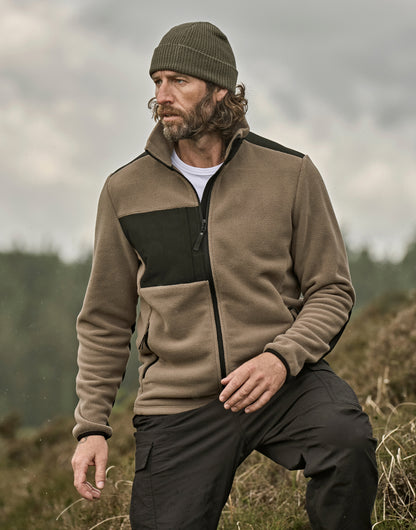 Mann trägt eine Mountain Fleecejacke in Clay/Black mit schwarzem Reißverschluss und dunklen Kontrastbesätzen an Brust, Schultern und Ärmeln. Die hüftlange Jacke ist körpernah geschnitten, hat einen hohen Kragen und seitliche Reißverschlusstaschen.