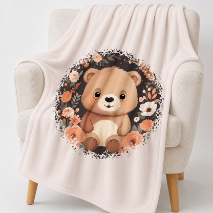 „Hellbeige Kuscheldecke mit einem sitzenden braunen Teddybären in der Mitte, umgeben von einem kreisförmigen Kranz aus dunklen Zweigen und Blüten in Creme-, Beige- und Apricottönen.“