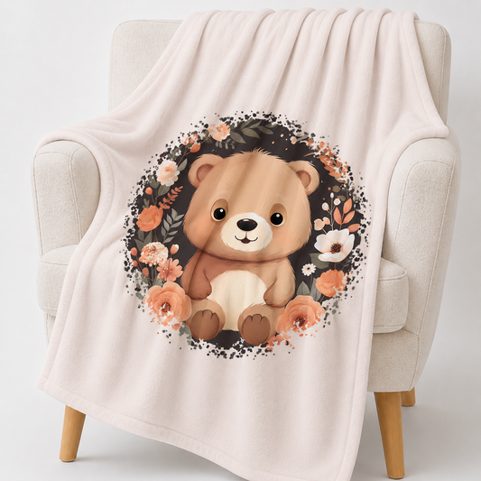 „Hellbeige Kuscheldecke mit einem sitzenden braunen Teddybären in der Mitte, umgeben von einem kreisförmigen Kranz aus dunklen Zweigen und Blüten in Creme-, Beige- und Apricottönen.“